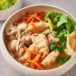 Lettuce, shredded carrots, wontons, chicken, and pork in broth that is in a white bowl.
