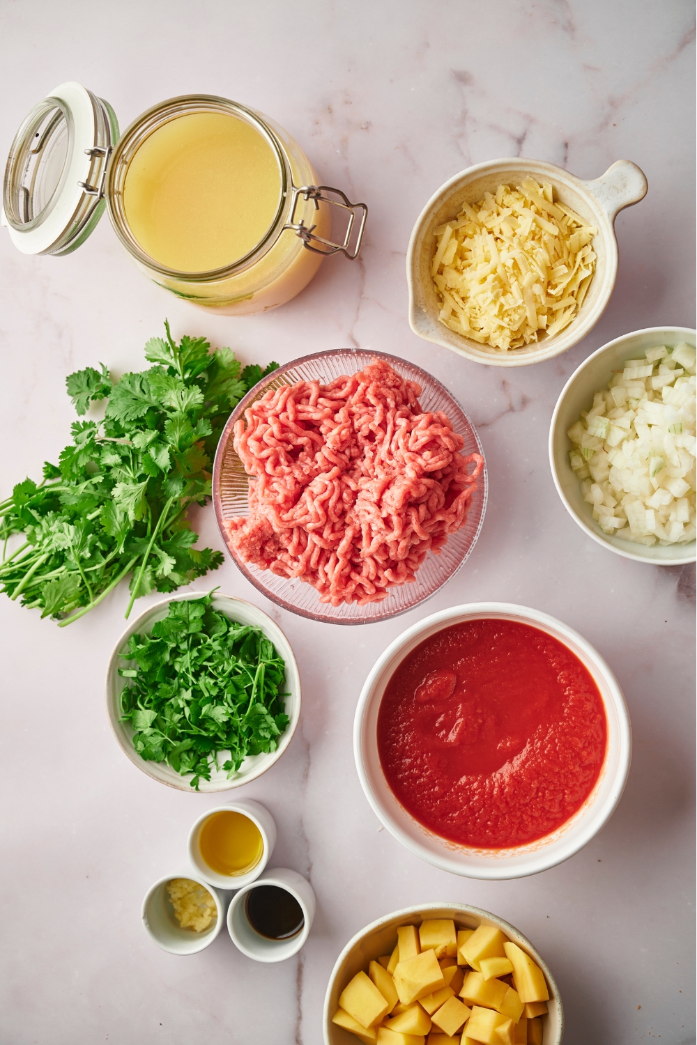 A bowl of ground beef, a glass jar of broth, a bowl of shredded cheese, a bowl of cilantro, a bowl of crushed tomatoes, and part of a bowl of potatoes.