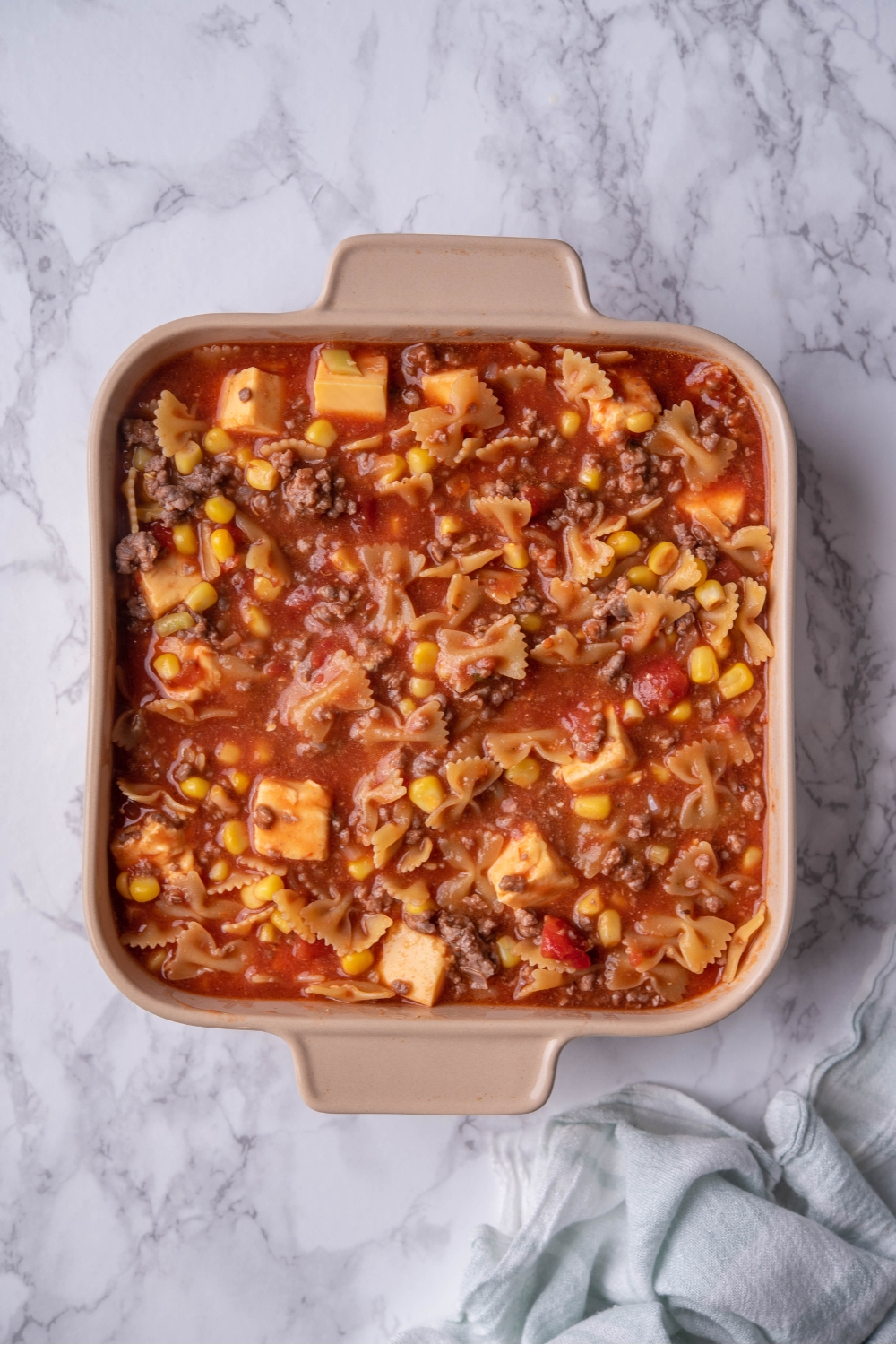 Bow tie pasta, corn, cheese cubes, and ground beef mixed in a sauce in a casserole dish.