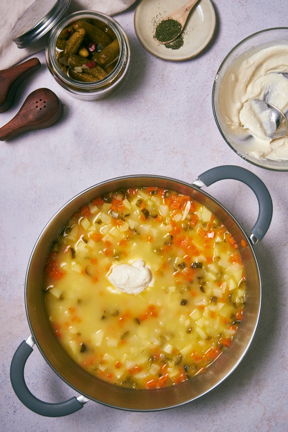 A dollop of sour cream in a pot that is filled with diced carrots, diced potatoes, diced pickles, and broth.