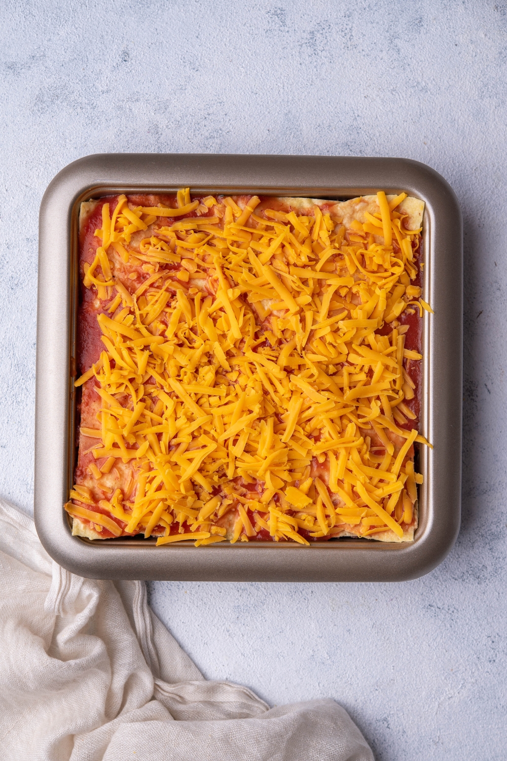 A bunch of shredded cheddar cheese on top of red enchilada sauce on an enchilada casserole and a baking dish.