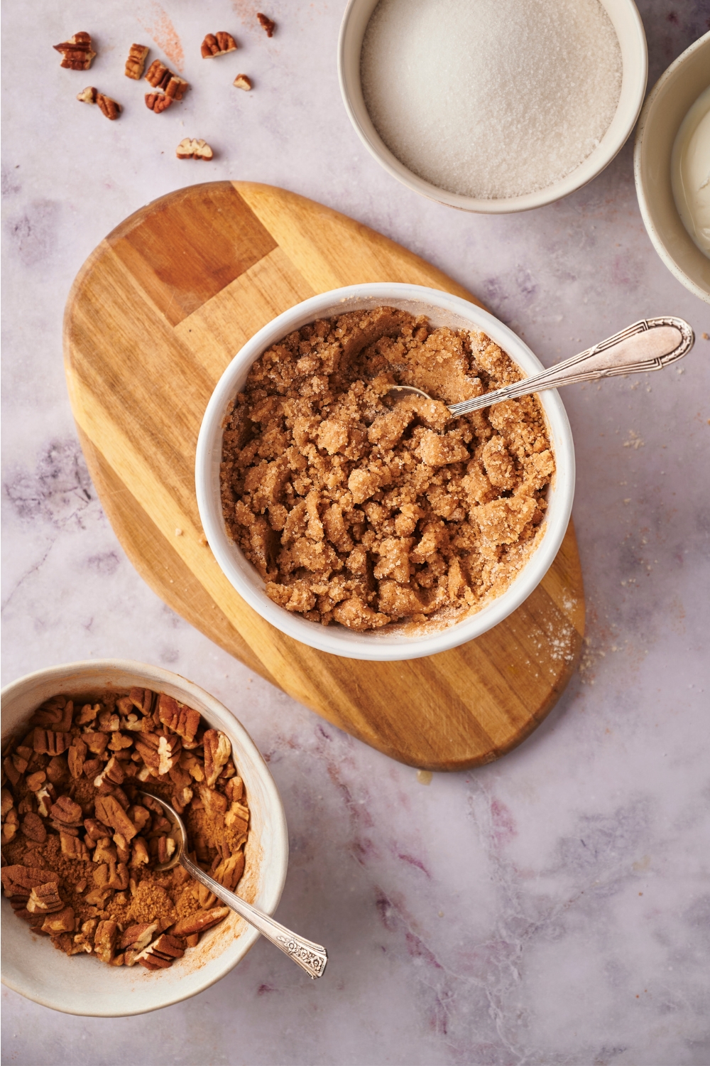 A brown sugar crumble mixture in a white bowl on top of a cutting board. In front of it is a pecan mixture in a white bowl.
