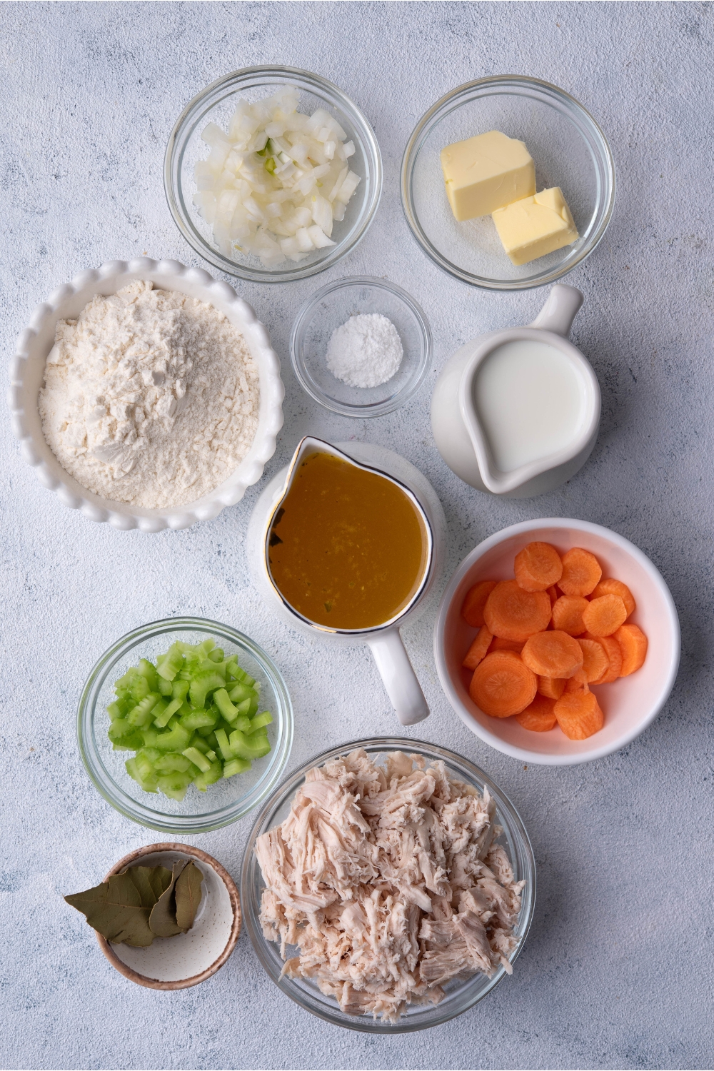 A bowl of onion, a bowl of butter, a bowl of flour, a bowl of salt, a pitcher of milk, a pitcher of broth, a bowl of celery, a bowl of carrots, a bowl of shredded chicken, and a bowl with a bay leaf in it all on a grey counter.