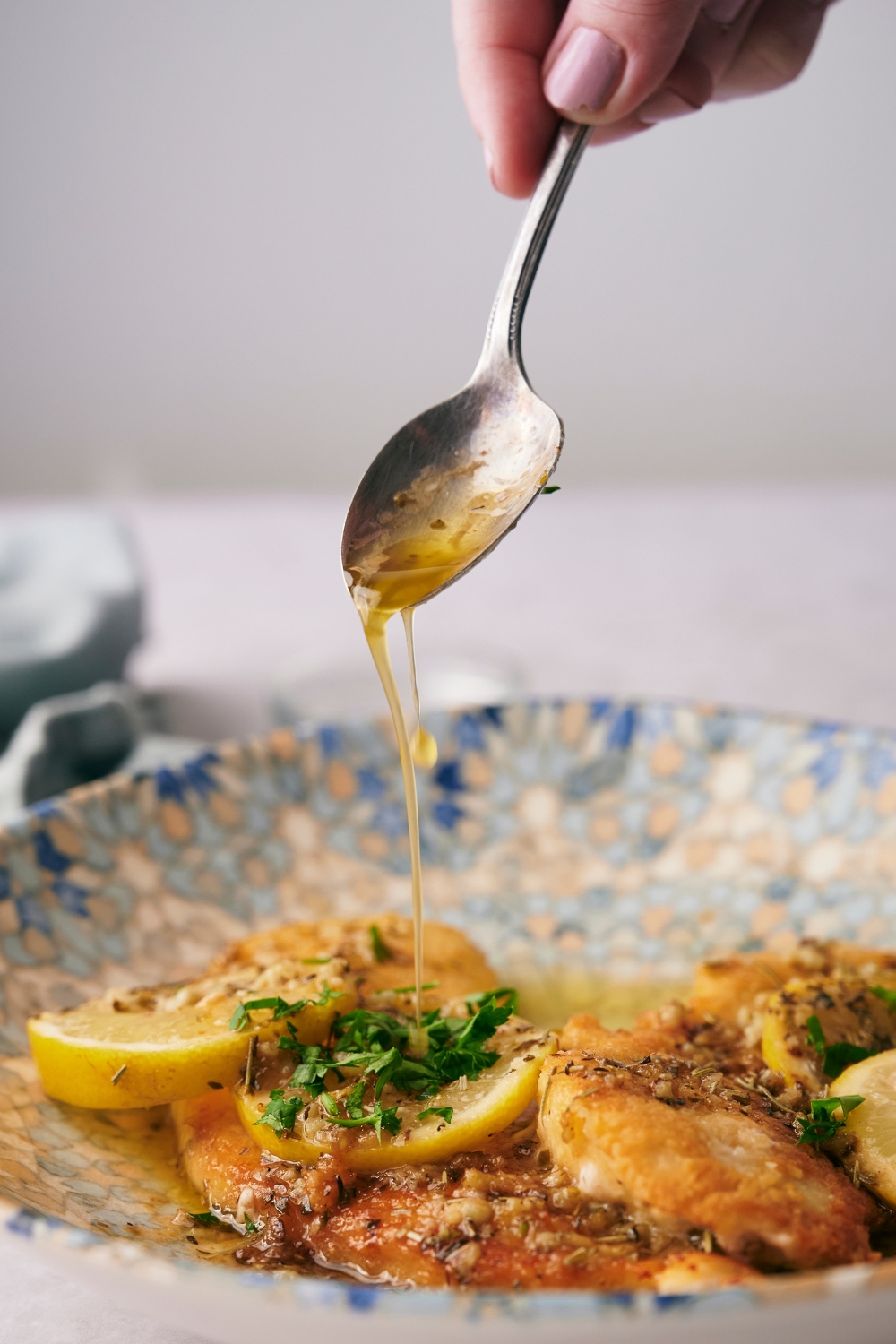 A plate with lemon chicken garnished with lemon and parsley. A spoon is drizzling butter lemon sauce over it.