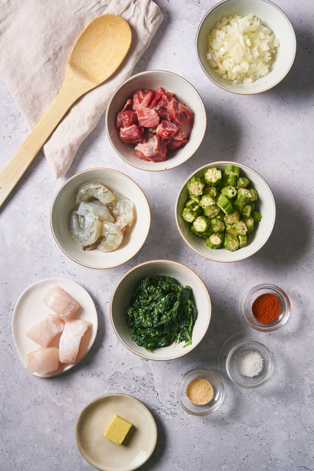 A bowl of onions, a bowl of cubed steak, a bowl of okra, a bowl of spinach, a plate with white fish on it, and a few bowls of seasonings all on a grey counter.