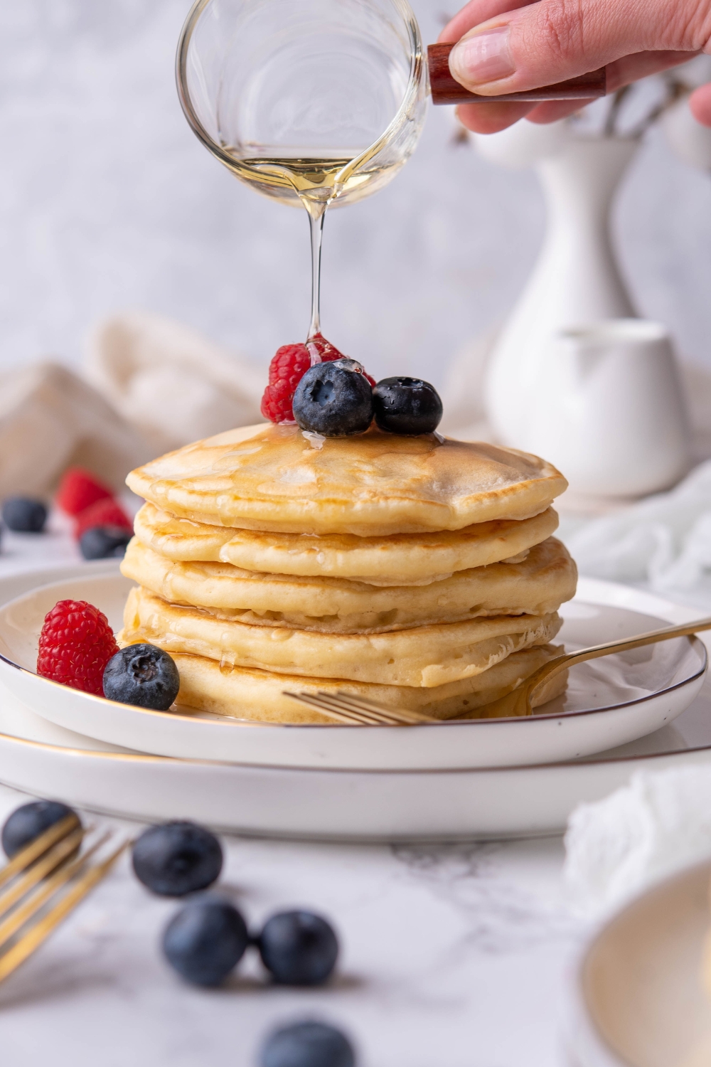 A stack of pancakes being drizzled with maple syrup and topped with fresh berries on a plate.