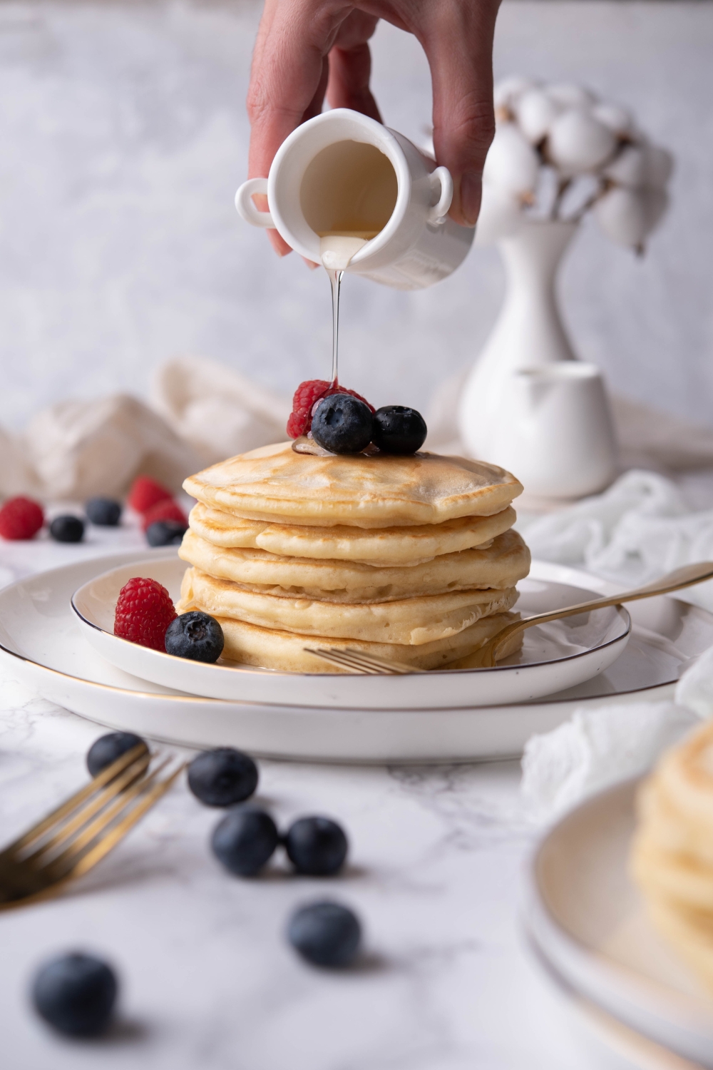A stack of pancakes being drizzled with maple syrup and garnished with fresh berries on a plate.