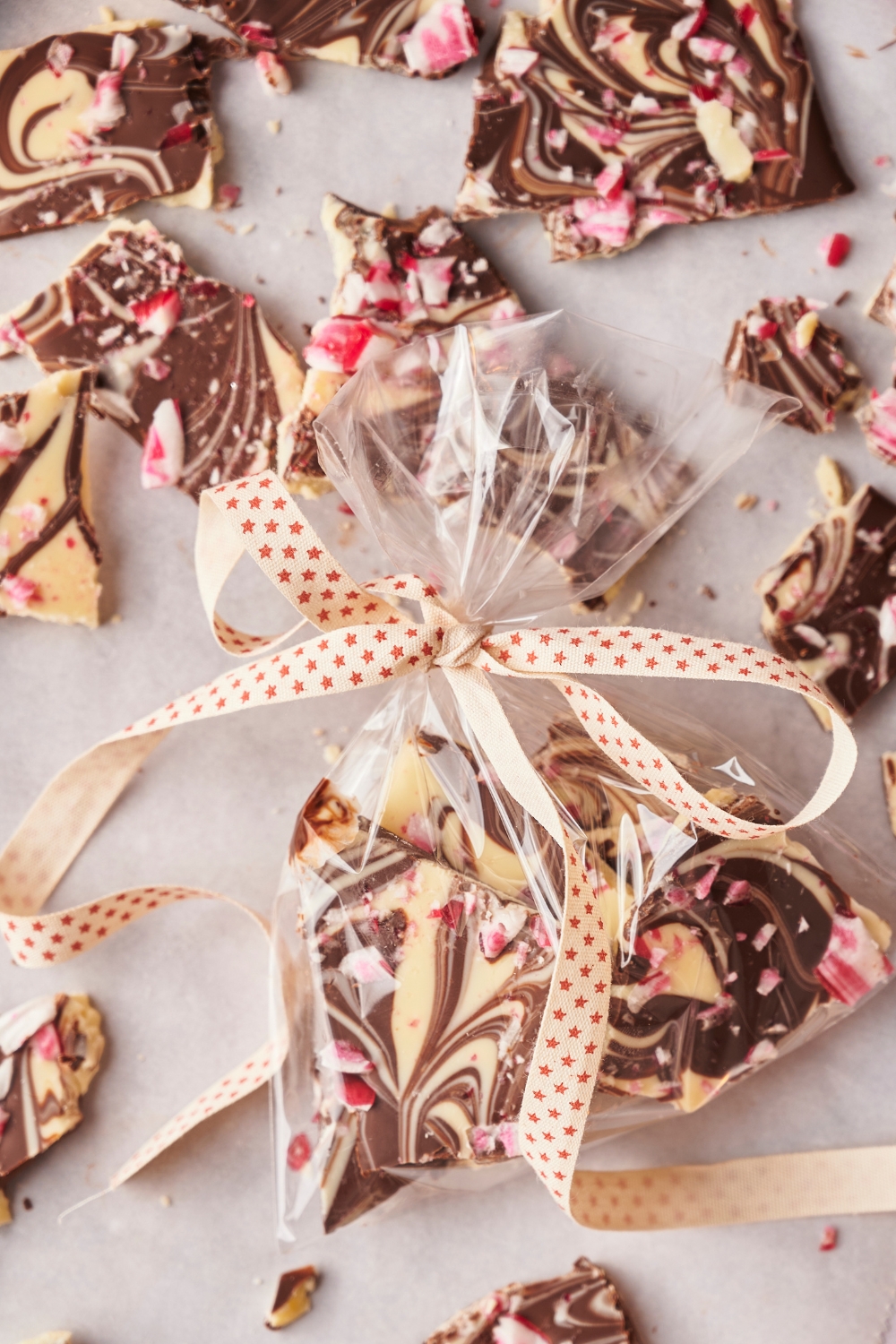 Peppermint bark pieces on parchment paper. A small clear bag of peppermint bark is tied with a ribbon and sitting in the middle of the peppermint bark pieces.