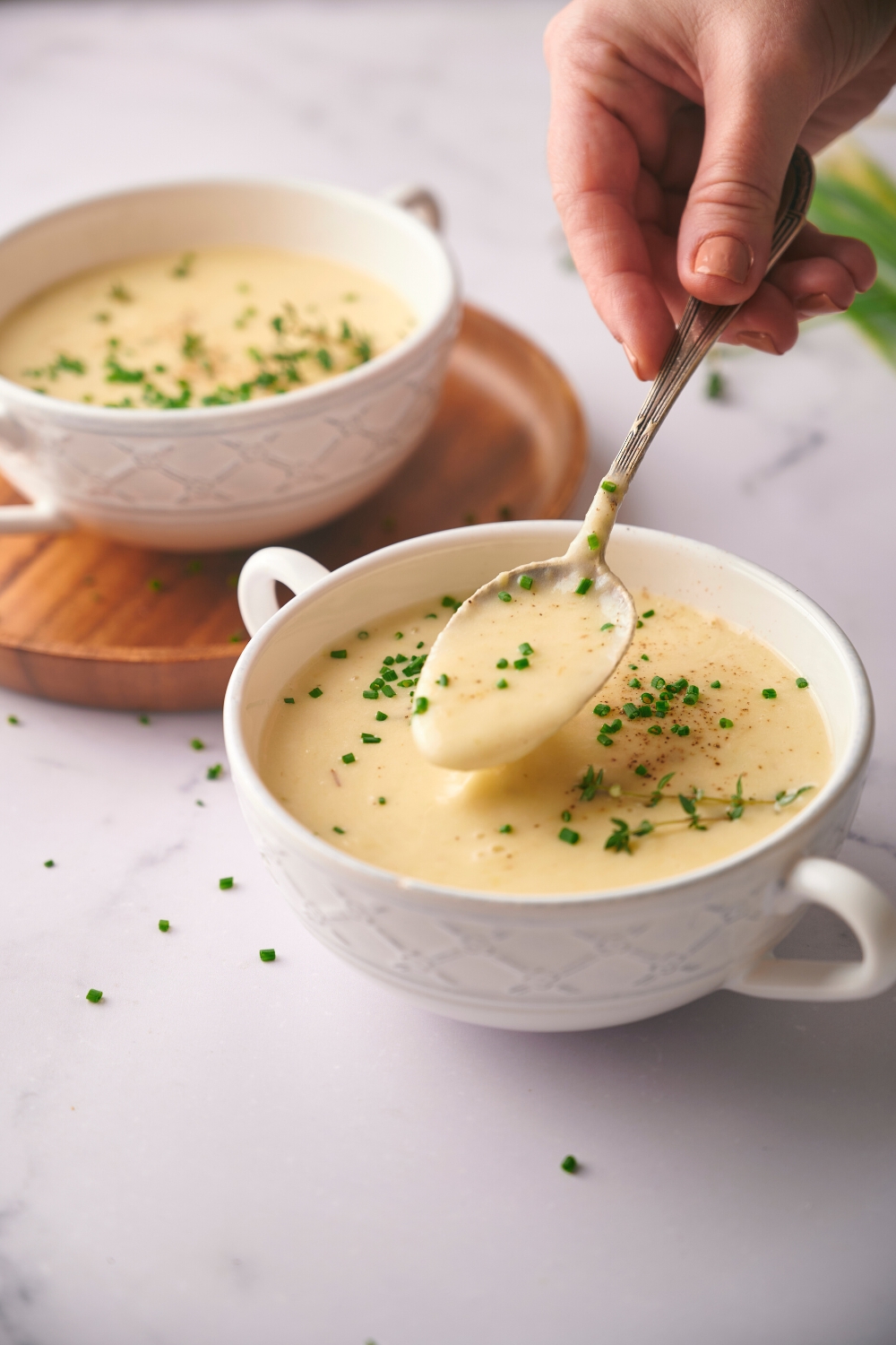A hand holding a spoon with potato leek soup on it directly above a white bowl that is filled with the soup. Behind it is another bowl of creamy soup on a circular wooden board.