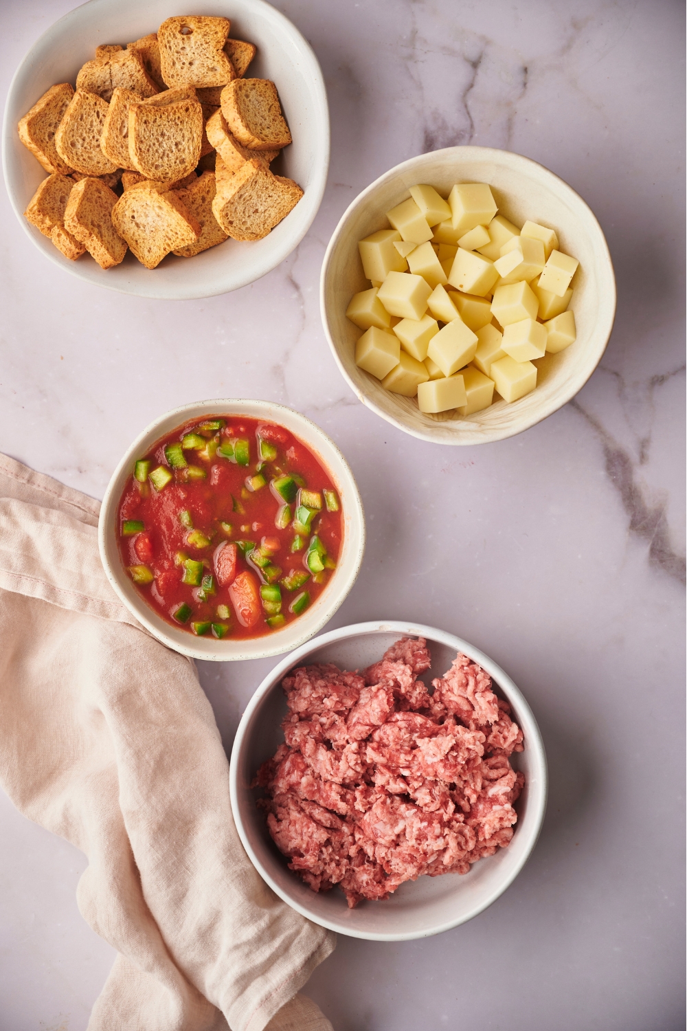 A bowl of mini toasts, a bowl of cubed cheese, a bowl of rotel tomatoes, and a bowl of ground sausage.