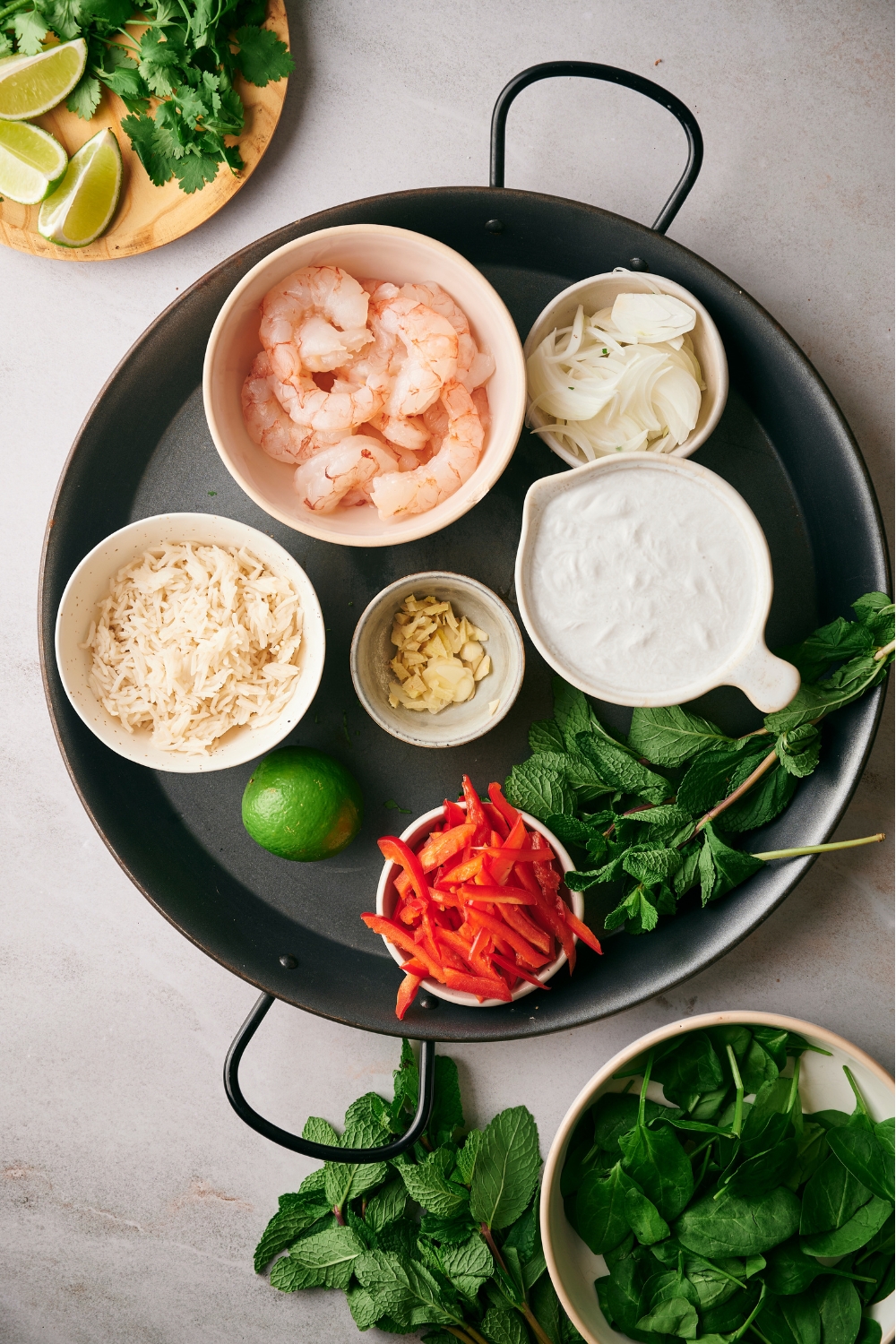 A bowl of shrimp, a bowl of diced onion, a bowl of rice, a bowl of ginger, a bowl of sliced bell peppers, and a lime on a serving plate.