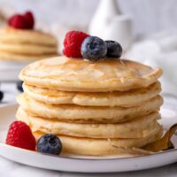 A stack of five pancakes with blueberries and a raspberry on top on a plate.