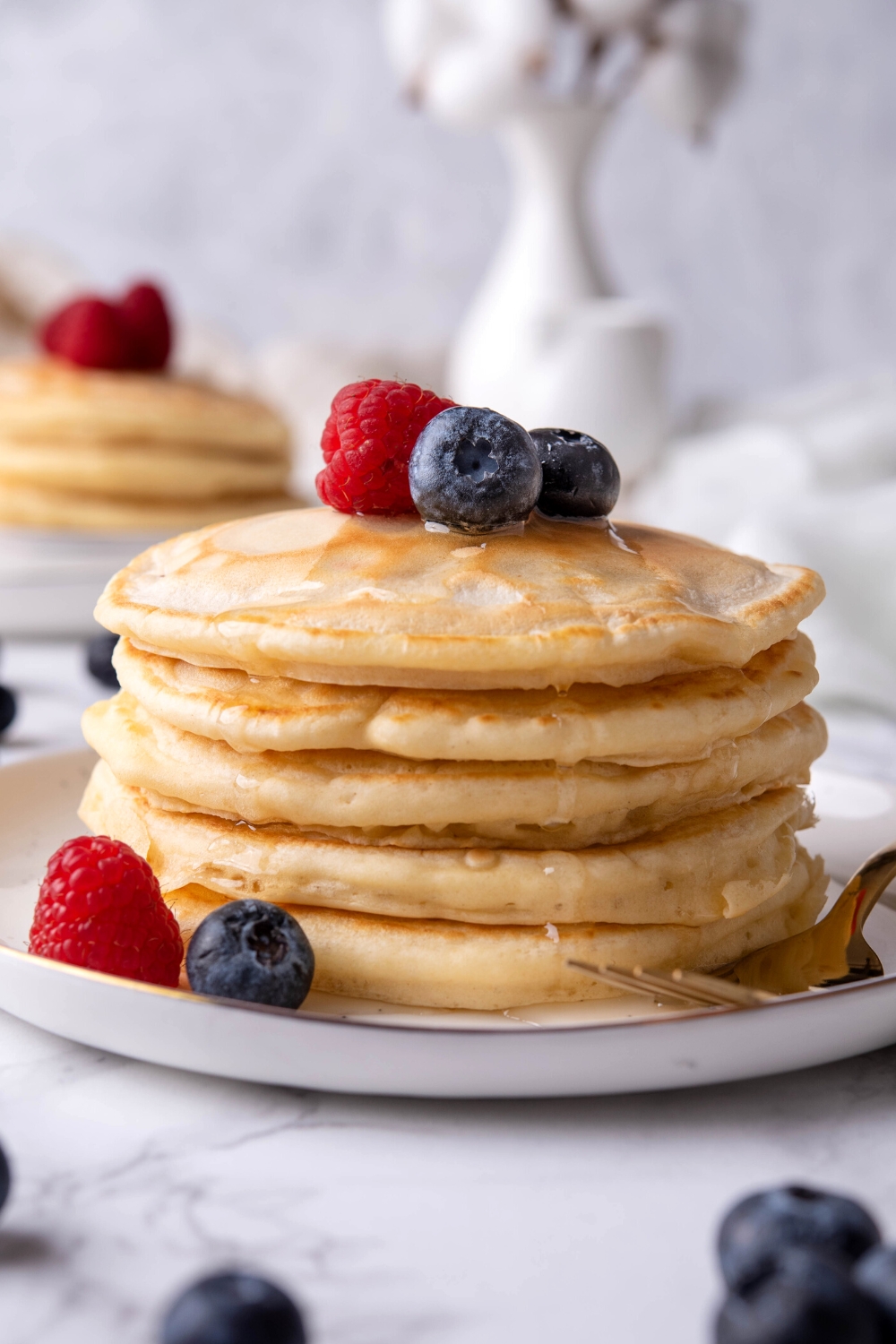 A stack of five pancakes with blueberries and a raspberry on top on a plate.