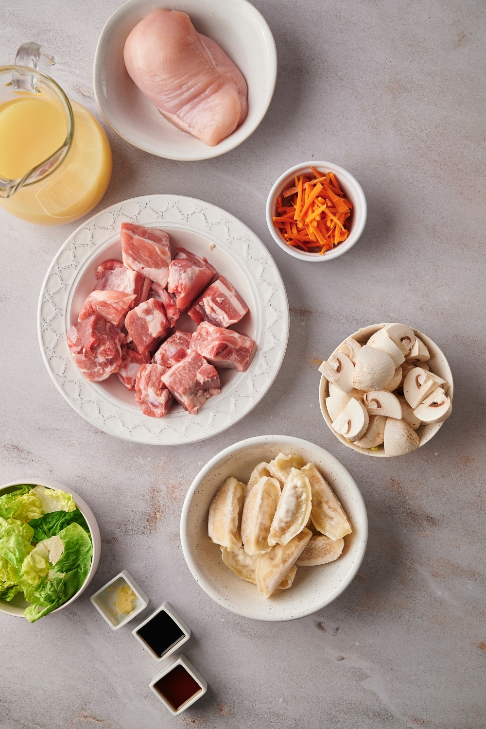 Bowl of cubed pork, a bowl of carrots, bowl with a chicken breast in it, a bowl of wontons, a bowl of soy sauce, a bowl of garlic, a bowl of lettuce, and a bowl of sesame oil all on a gray counter.
