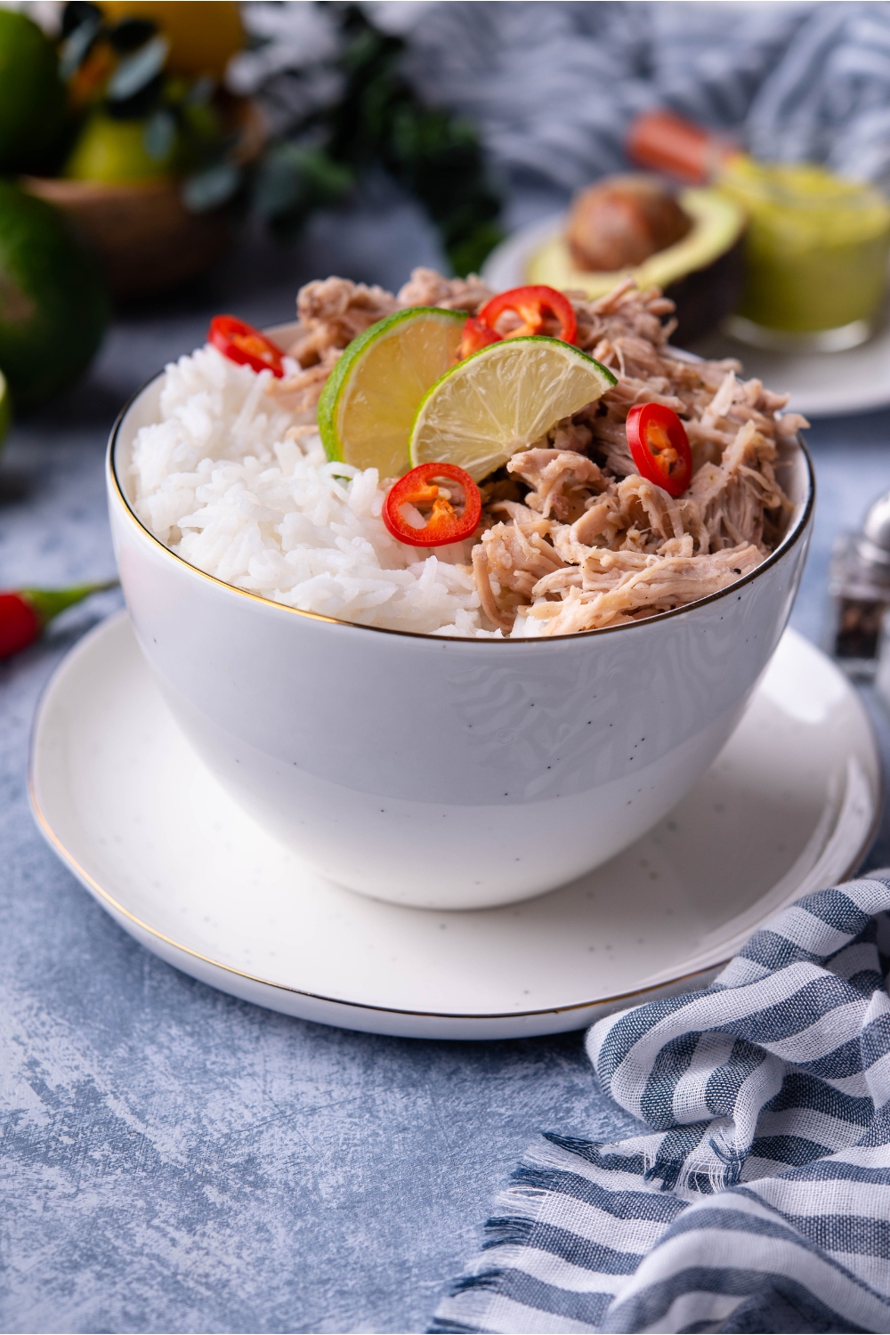 Shredded carnitas in a white bowl atop a white plate, with white rice, lime wedges, and sliced peppers.