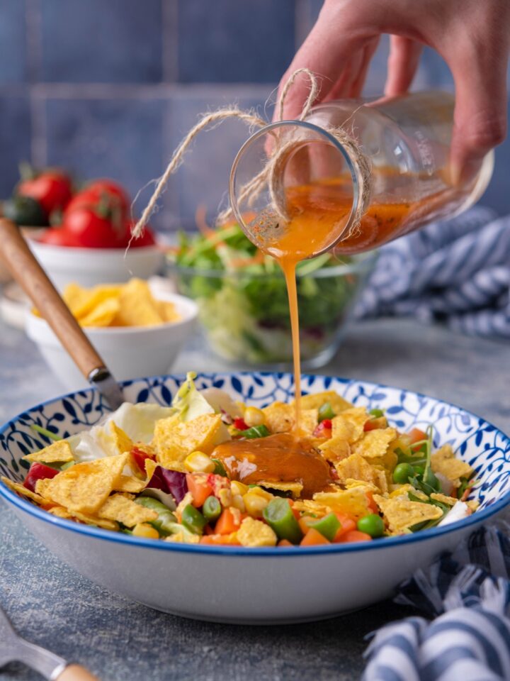 Hand drizzling vinaigrette over a salad of lettuce, tortilla chips, and assorted veggies.