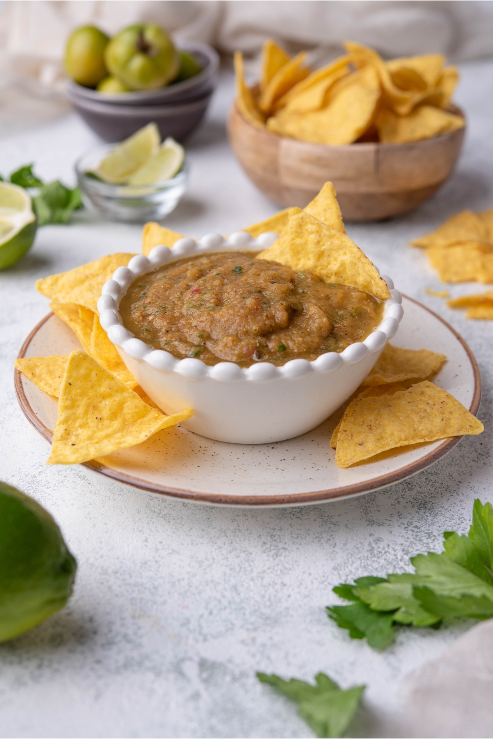 Tomatillo salsa in a small white bowl with tortilla chips in the salsa. The bowl is on top of a plate filled with tortilla chips. In the background there is a bowl of tomatillos and a bowl of tortilla chips.