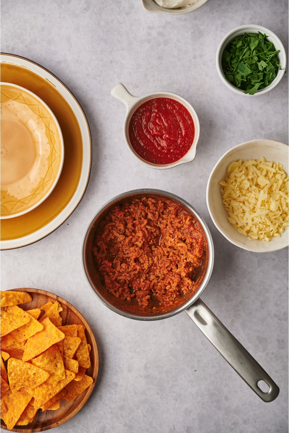 A sauce pot filled with chili surrounded by bowls of shredded cheese, fresh herbs, salsa, and tortilla chips.