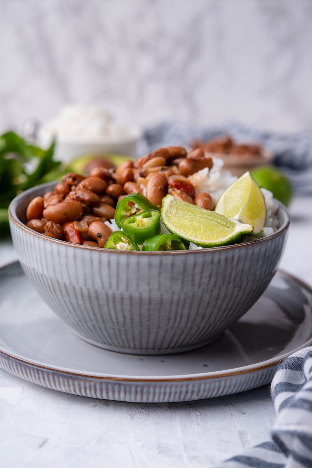 A grey bowl atop a grey plate filled with pinto beans and white rice, garnished with lime wedges and sliced peppers.