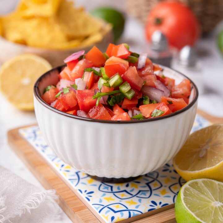 Fresh tomato salsa in a white bowl atop a decorative serving tray, surrounded by limes and lemons. There are bowls of tomatoes and tortilla chips in the background.