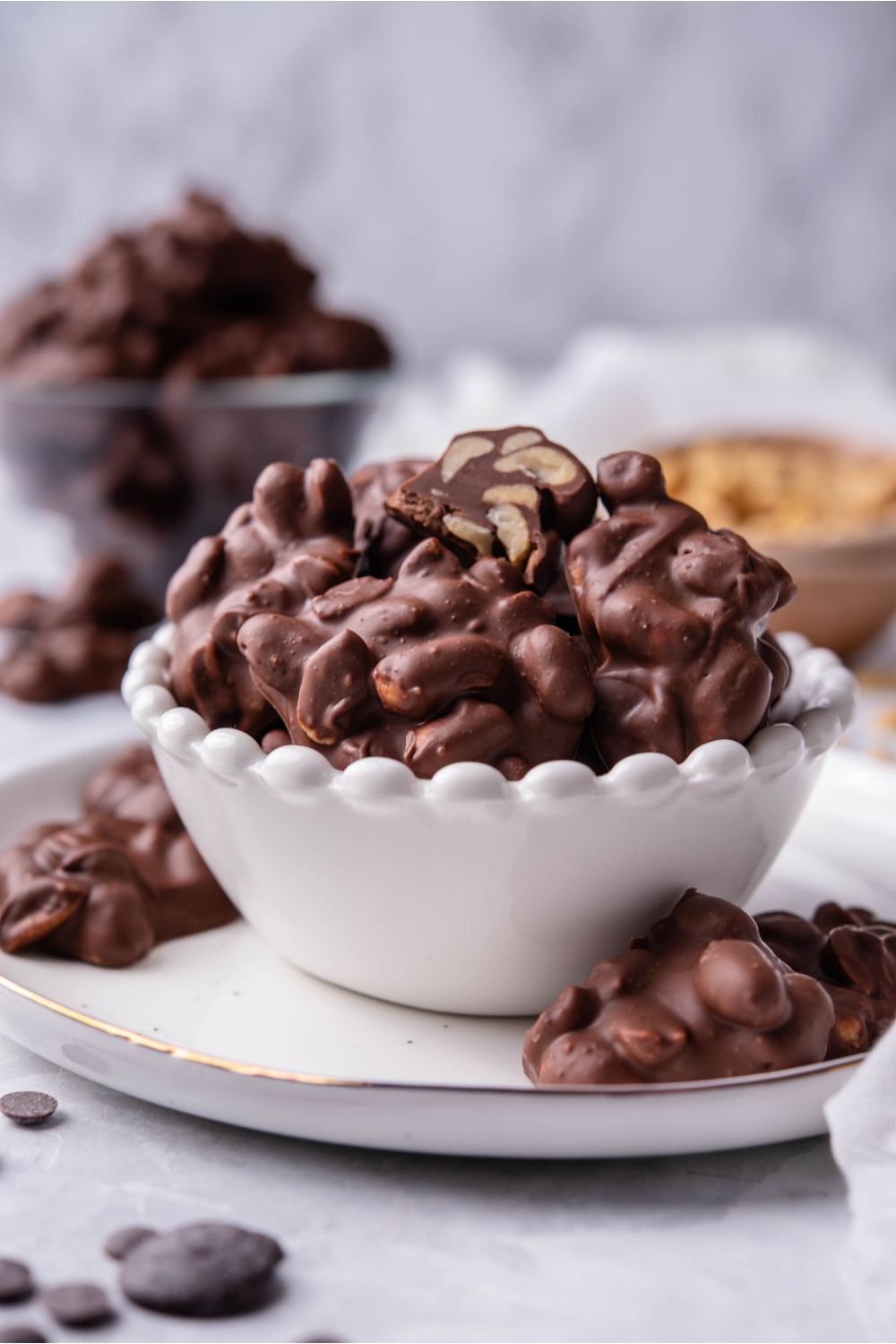 Crock pot chocolate peanut clusters piled in a white serving bowl with a one cluster cut in half and some pieces spilling out on the plate below. In the background is another bowl of peanut clusters and a bowl of peanuts.