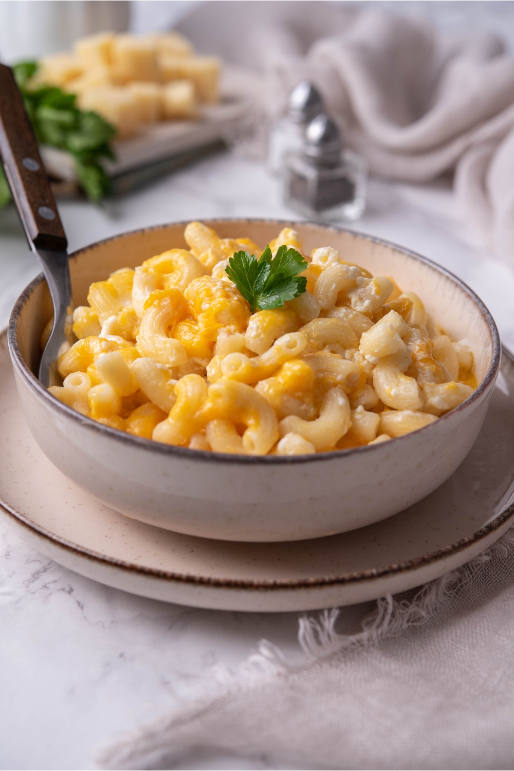 Old-fashioned macaroni and cheese in a white bowl with a fork and a garnish of fresh parsley in the bowl. The bowl is atop a white plate.