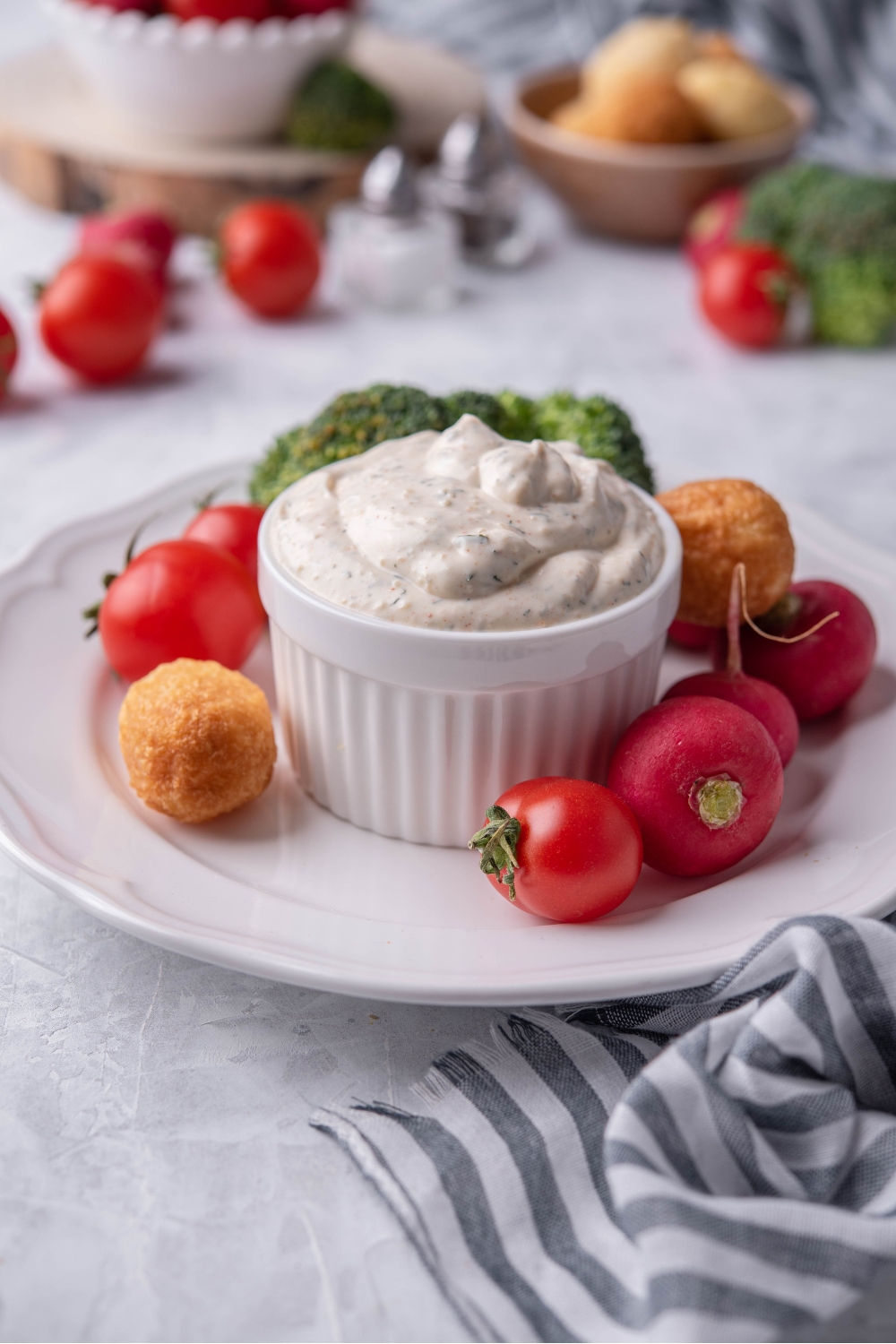 Vegetable dip in a white ramekin atop a white plate that is filled with cherry tomatoes, broccoli, and nuggets. In the background are bowls of tomatoes, nuggets, and broccoli.