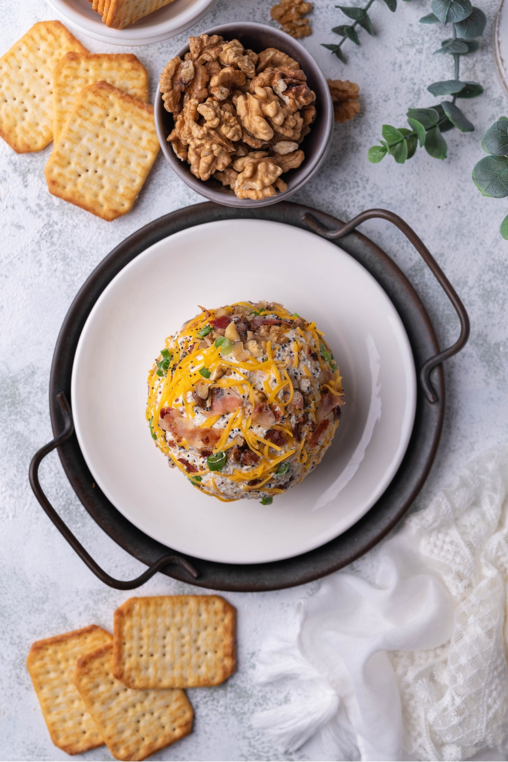 Overview of a cheese ball on a white plate layered with a serving tray. The cheese ball is surrounded by crackers and a bowl of chopped nuts.