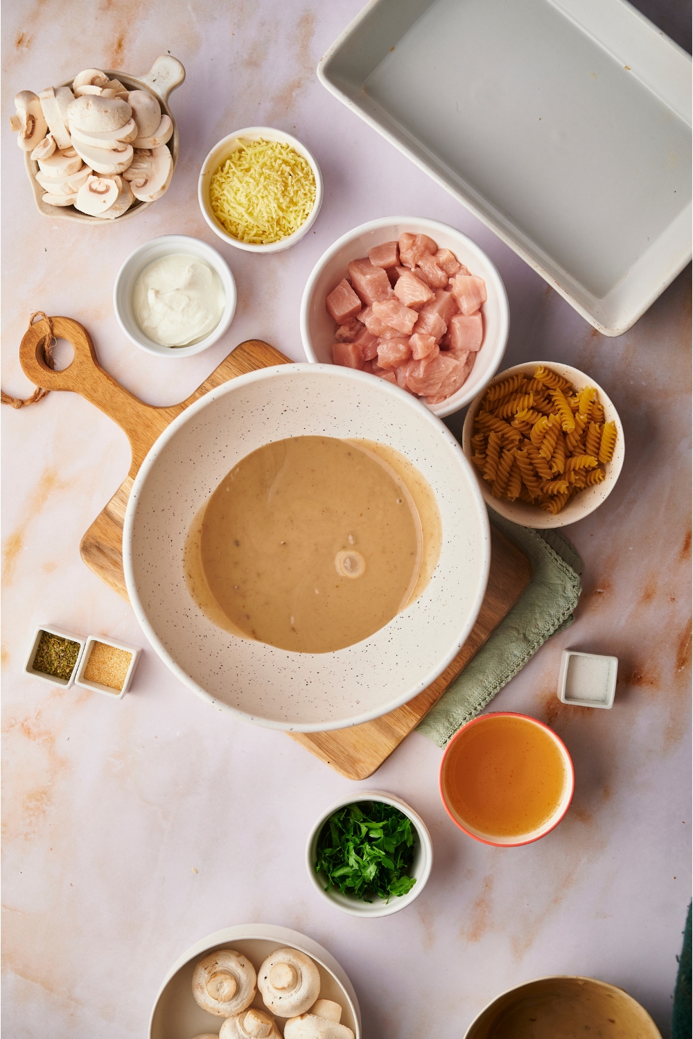 A white bowl filled with a creamy soup mixture and surrounded by ingredients including bowls of raw chicken, dried pasta, broth, raw sliced mushrooms, spices, and sour cream.