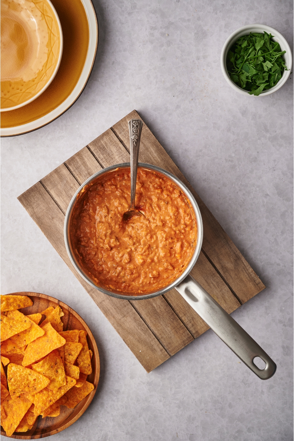 A sauce pan filled with chili cheese dip on top of a wooden board. There is a spoon in the pot and the pot is next to a bowl of fresh herbs and a plate of tortilla chips.