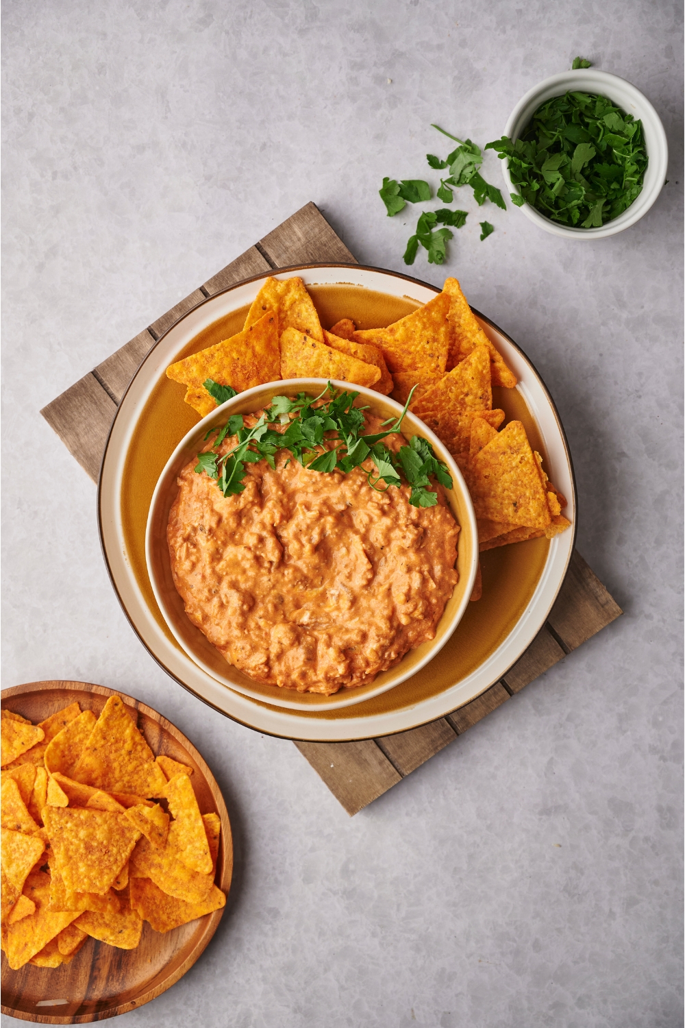 Chili cheese dip in a serving bowl atop a plate layered with tortilla chips, next to a bowl of fresh herbs and a plate of more tortilla chips.