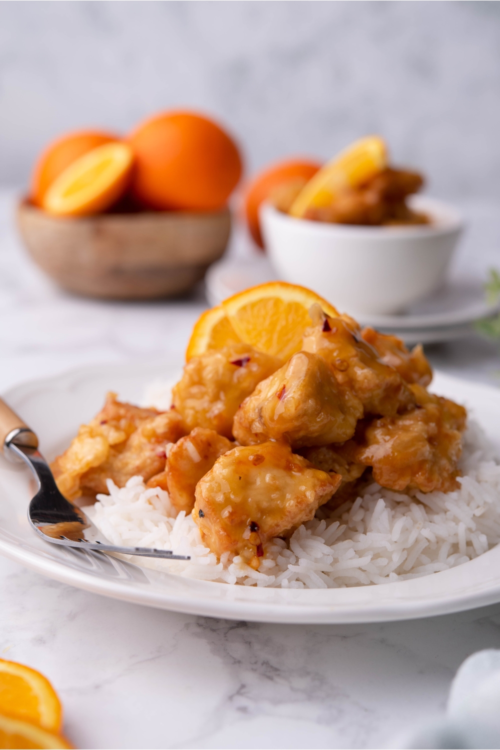 Orange chicken on a bed of white rice on a white plate with an orange slice and a fork on the plate.