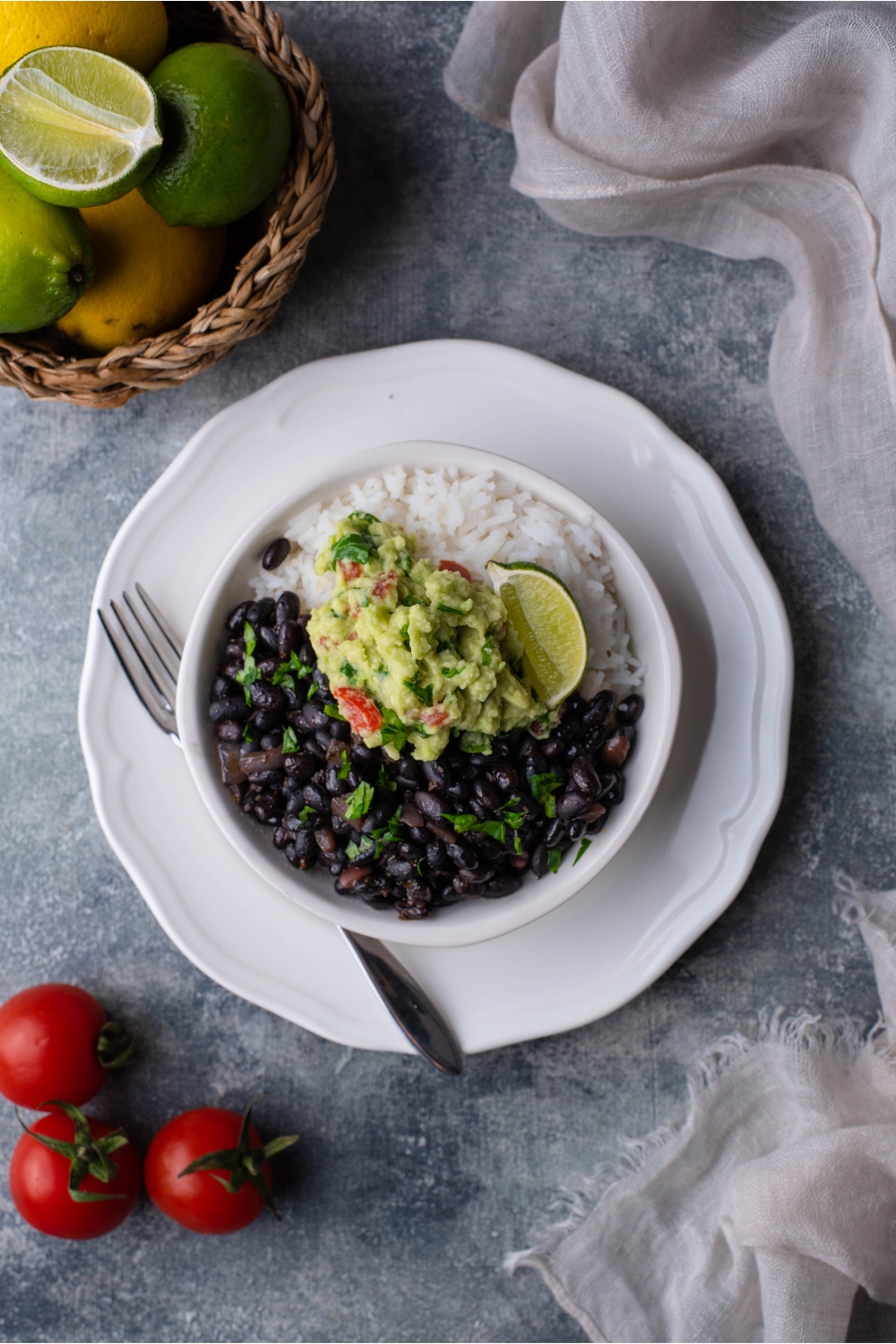 Overview of Chipotle black beans in a white bowl layered on a white plate, served with white rice, guacamole, a lime wedge, and fresh herbs. A fork is on the plate.