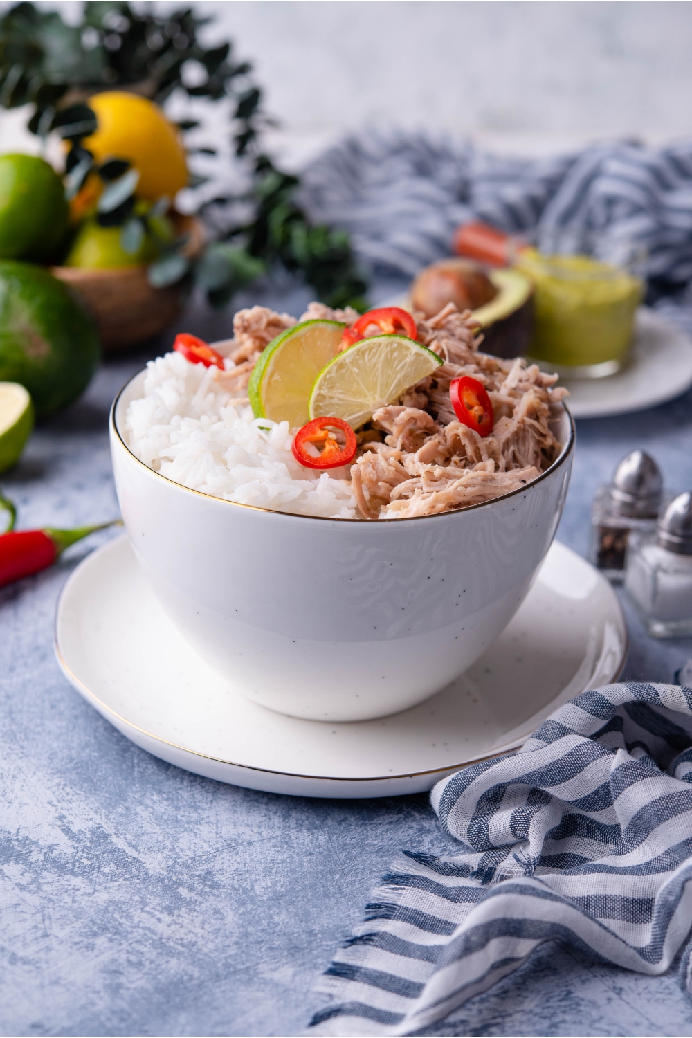 Shredded carnitas in a white bowl atop a white plate, with white rice, lime wedges, and sliced peppers.