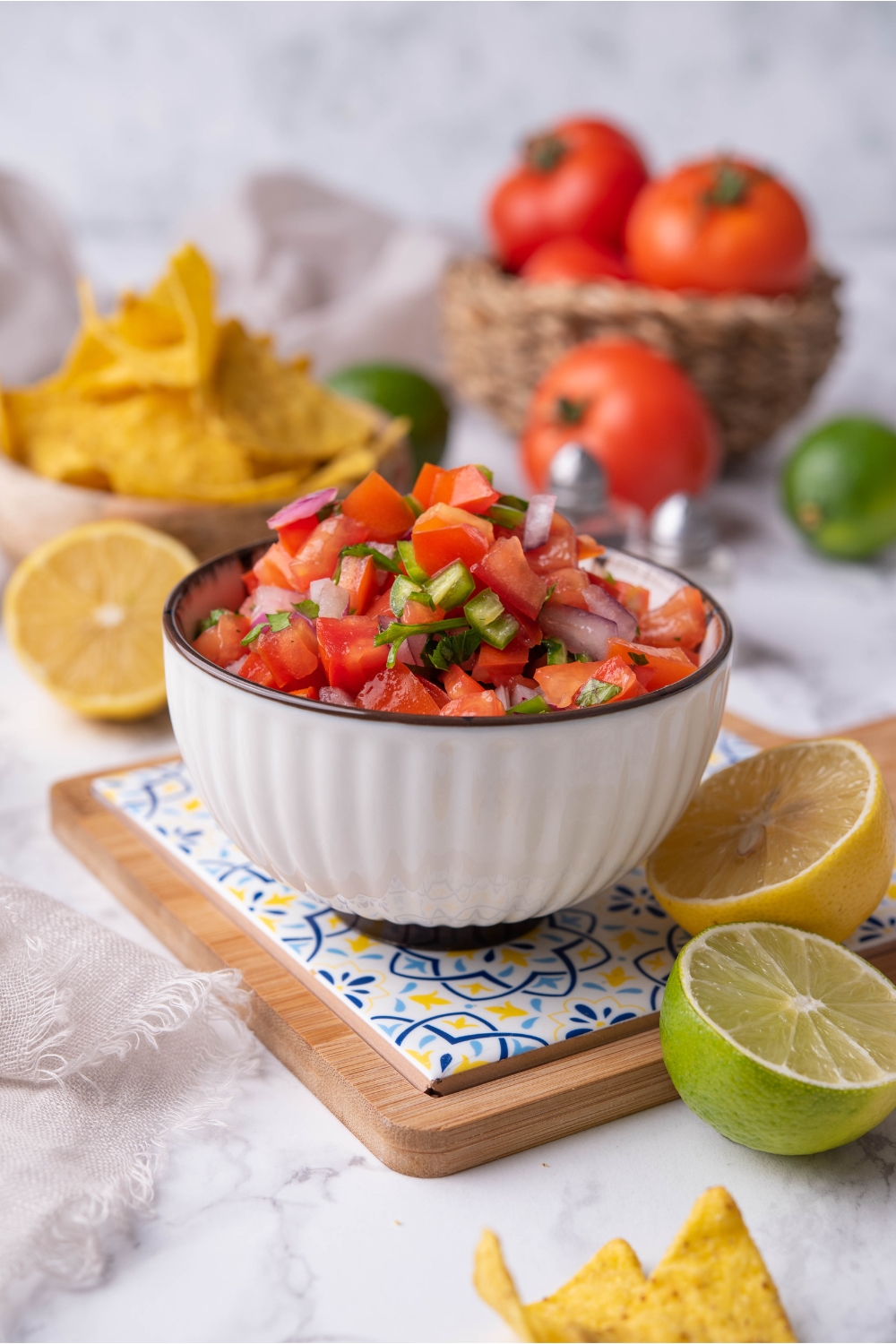 Fresh tomato salsa in a white bowl atop a decorative serving tray, surrounded by limes and lemons. There are bowls of tomatoes and tortilla chips in the background.