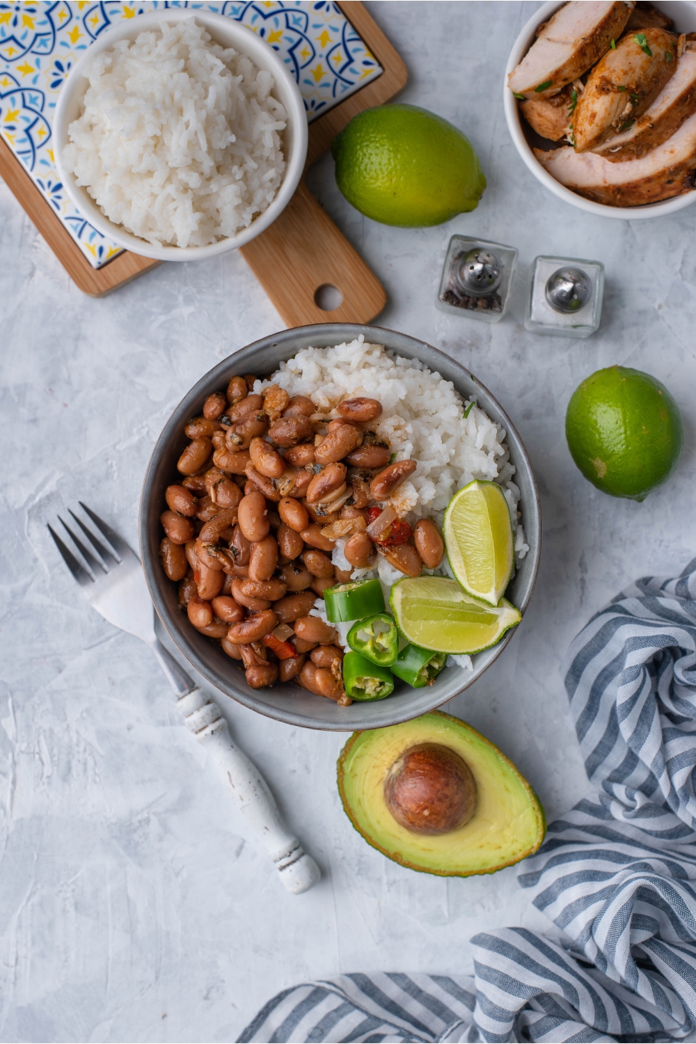 Overhead view of a bowl of pinto beans and white rice garnished with lime wedges and sliced peppers. The bowl is surrounded by a halved avocado, limes, a bowl of white rice, a fork, salt and pepper shakers, and sliced meat.
