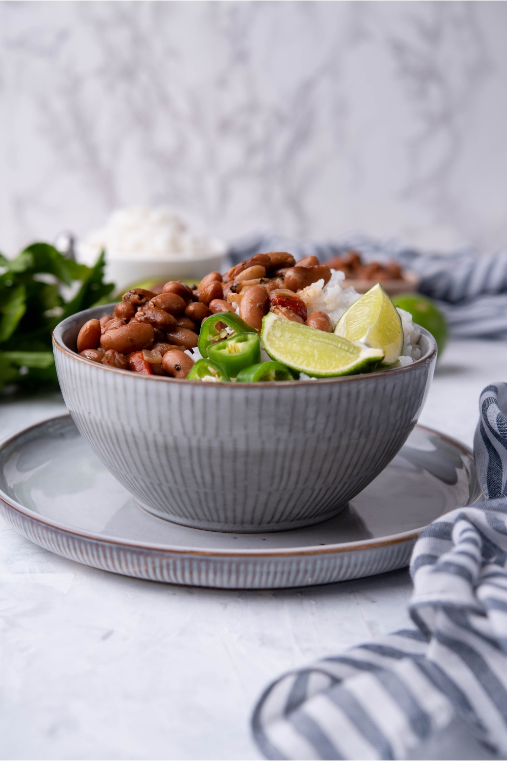 A grey bowl atop a grey plate filled with pinto beans and white rice, garnished with lime wedges and sliced peppers.