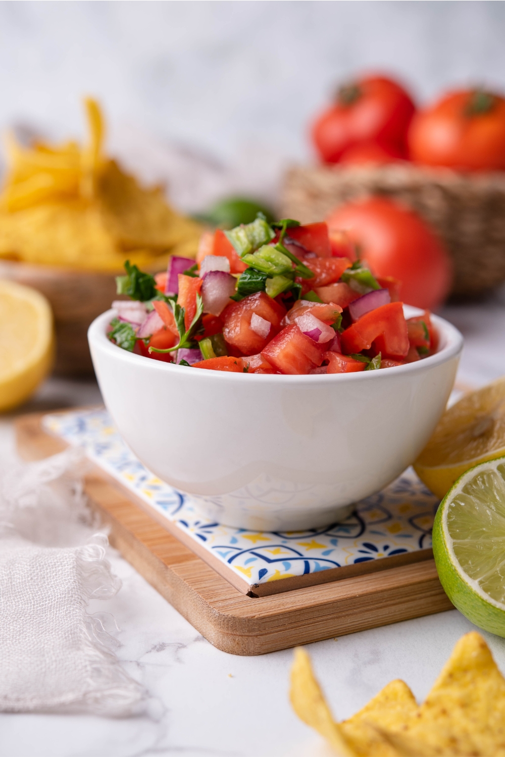 Fresh tomato salsa in a white bowl atop a decorative serving tray, surrounded by limes and lemons. There are bowls of tomatoes and tortilla chips in the background.