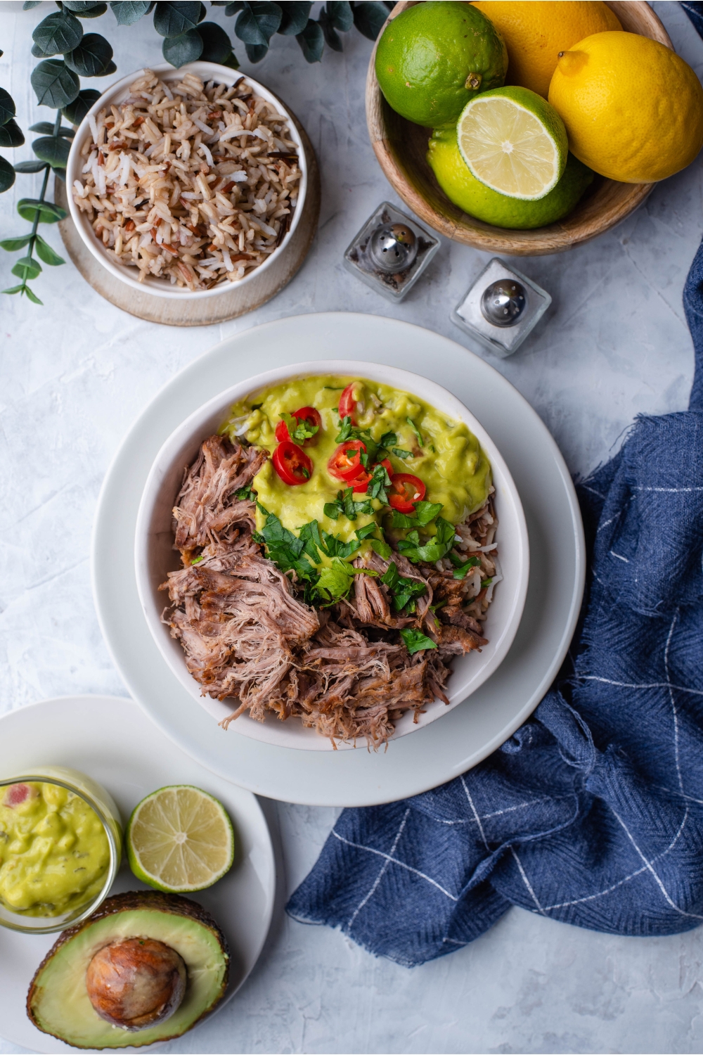 Overview of shredded barbacoa in a bowl with brown rice, sliced peppers, guacamole, and fresh herbs.