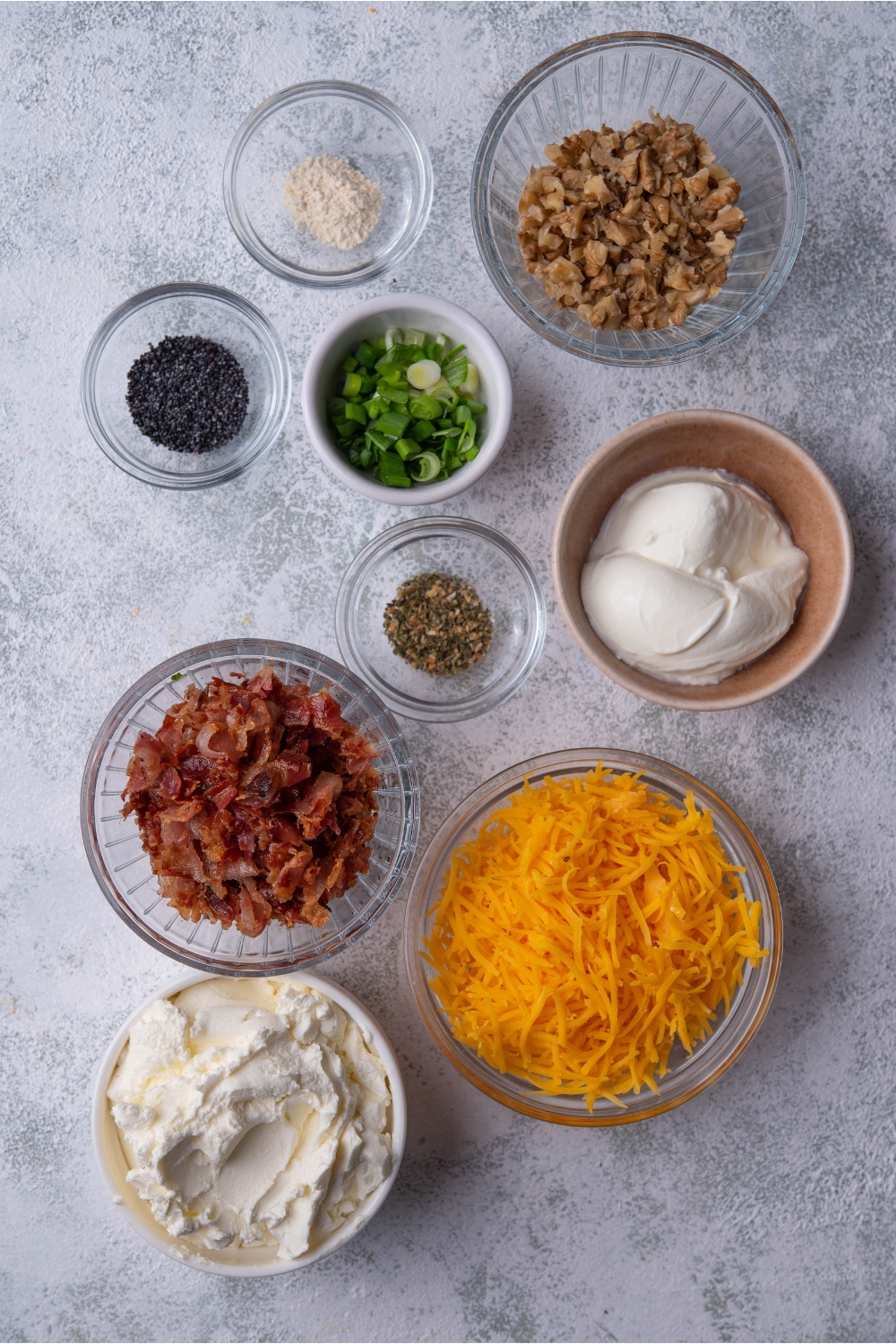 An assortment of ingredients including bowls of shredded cheese, cream cheese, chopped nuts, bacon, green onion, poppyseeds, and seasoning.