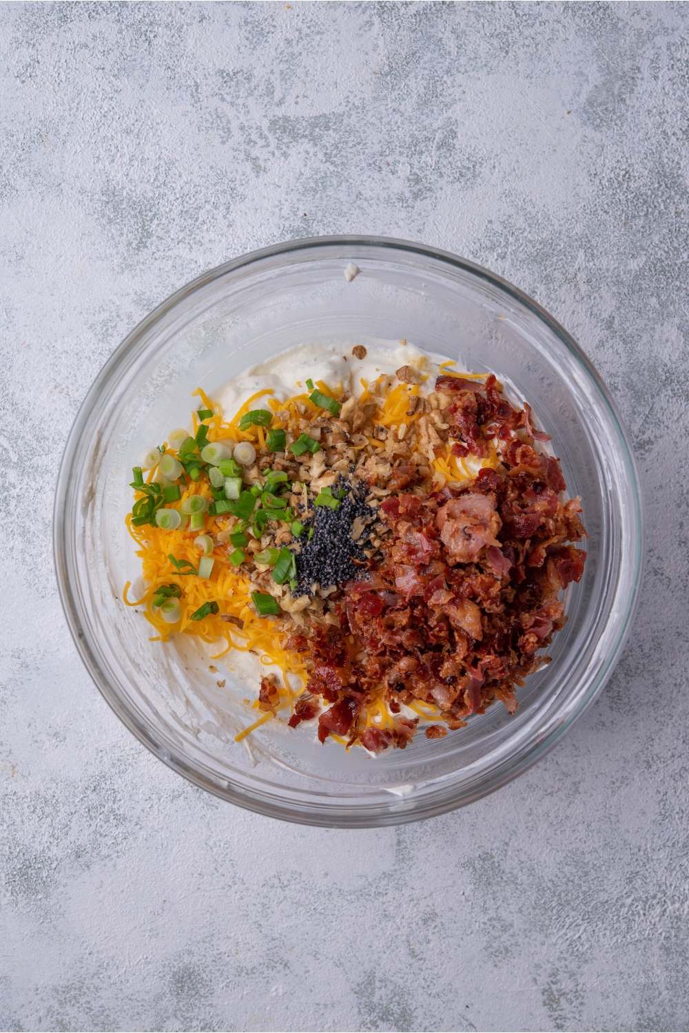 Clear bowl filled with a sour cream mixture, chopped bacon, shredded cheese, nuts, green onion, and poppyseeds.