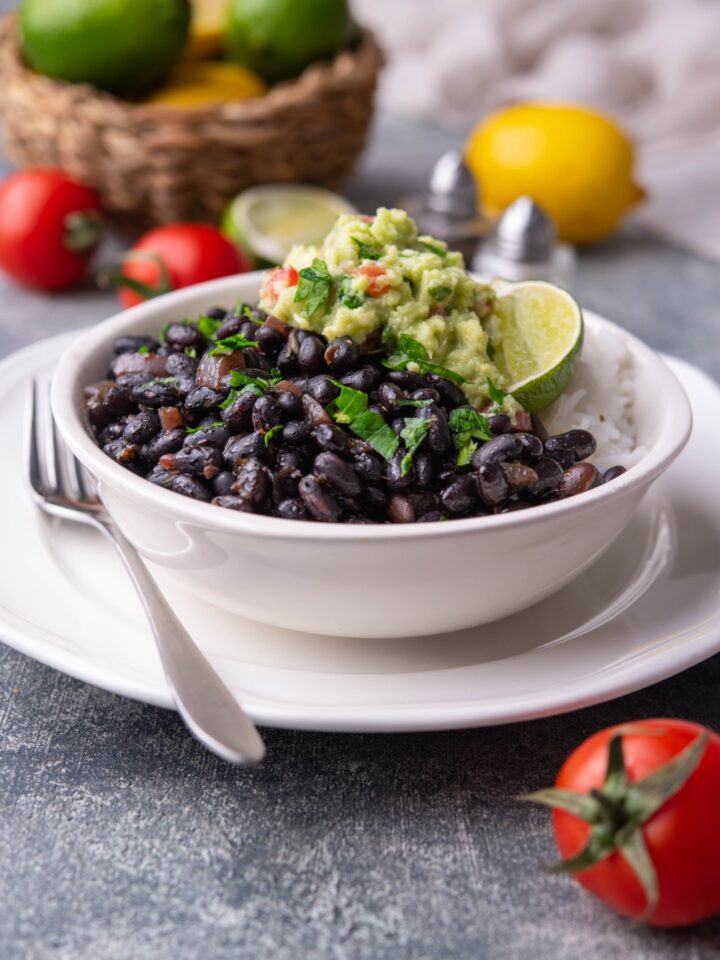 Chipotle black beans in a white bowl layered on a white plate, served with white rice, guacamole, a lime wedge, and fresh herbs. A fork is on the plate and a bowl of limes and lemons is in the background.