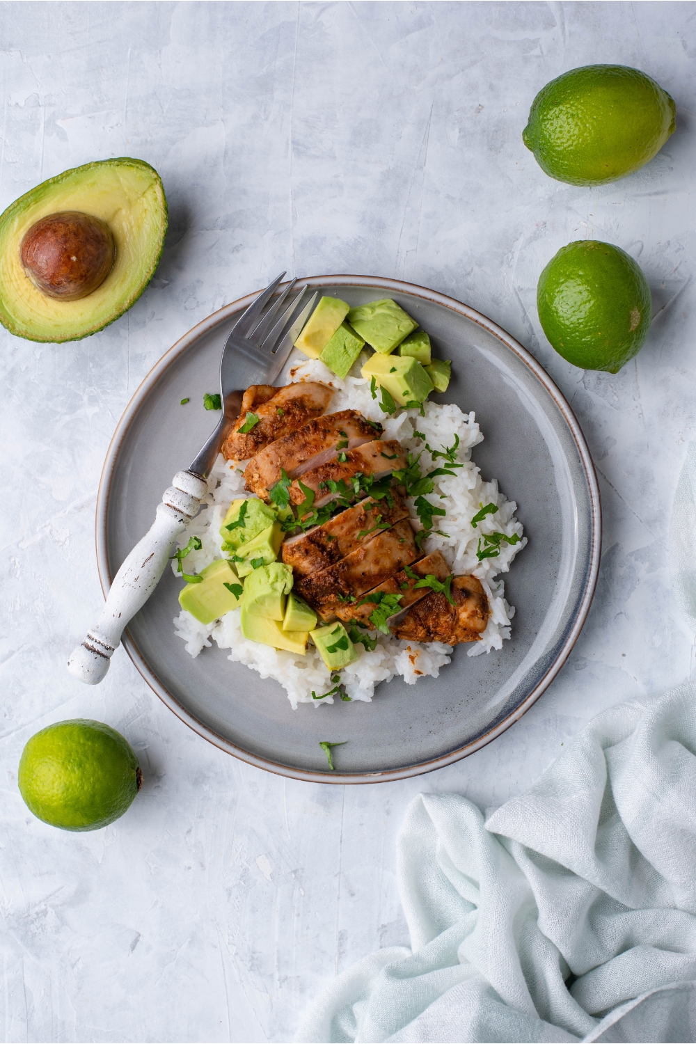 Overview of Chipotle chicken on a bed of white rice garnished with fresh herbs and diced avocado on a blue plate. The chicken is surrounded by limes and a halved avocado.