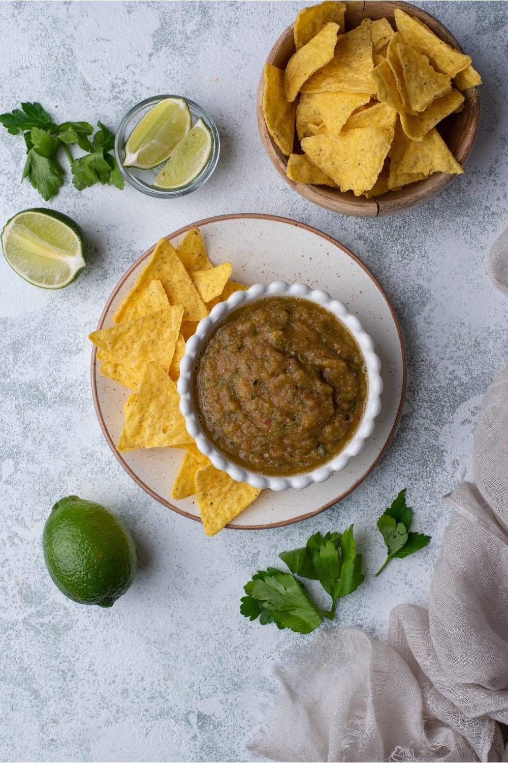 Tomatillo salsa in a small decorative bowl atop a plate filled with tortilla chips. Surrounding the plate of chips and salsa is a bowl of tortilla chips, a bowl of lime wedges, two limes, and fresh cilantro.