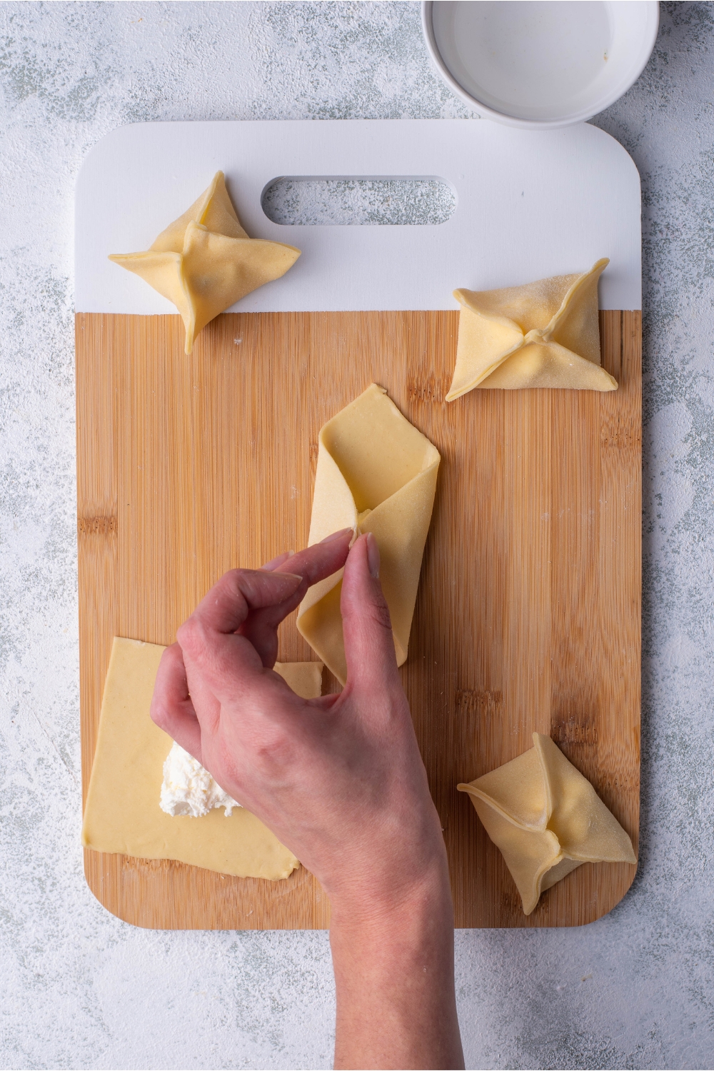 Hand pinching two corners of a wonton wrapper that is on a cutting board alongside cream cheese filled rangoon.