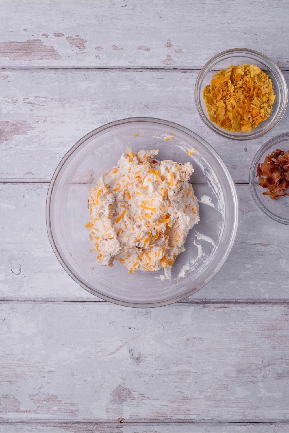 Clear bowl filled with a mixture of cream cheese, shredded cheese, and crumbled bacon. Next to the bowl are small bowls of bacon and crushed tortilla chips.