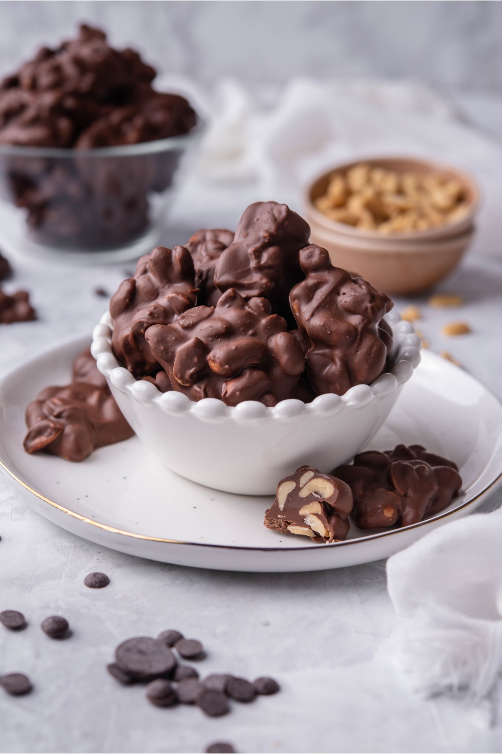 Crock pot chocolate peanut clusters piled in a white serving bowl with some pieces spilling out on the plate below. In the background is another bowl of peanut clusters and a bowl of peanuts.
