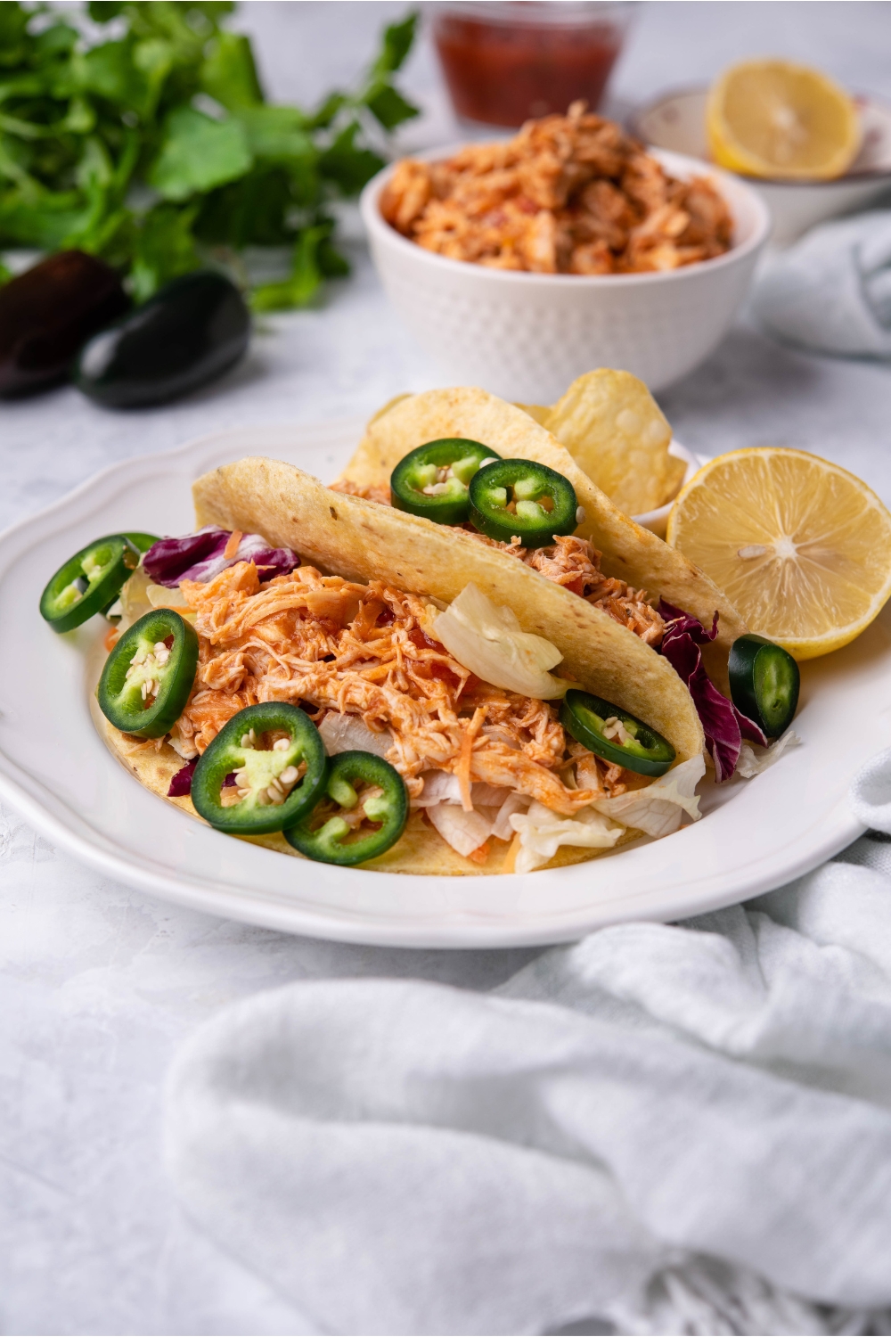Crockpot salsa chicken shredded in two tacos dressed with sliced jalapeños and shredded cabbage. There is a halved lemon on the plate and a bowl of shredded salsa chicken in the background.
