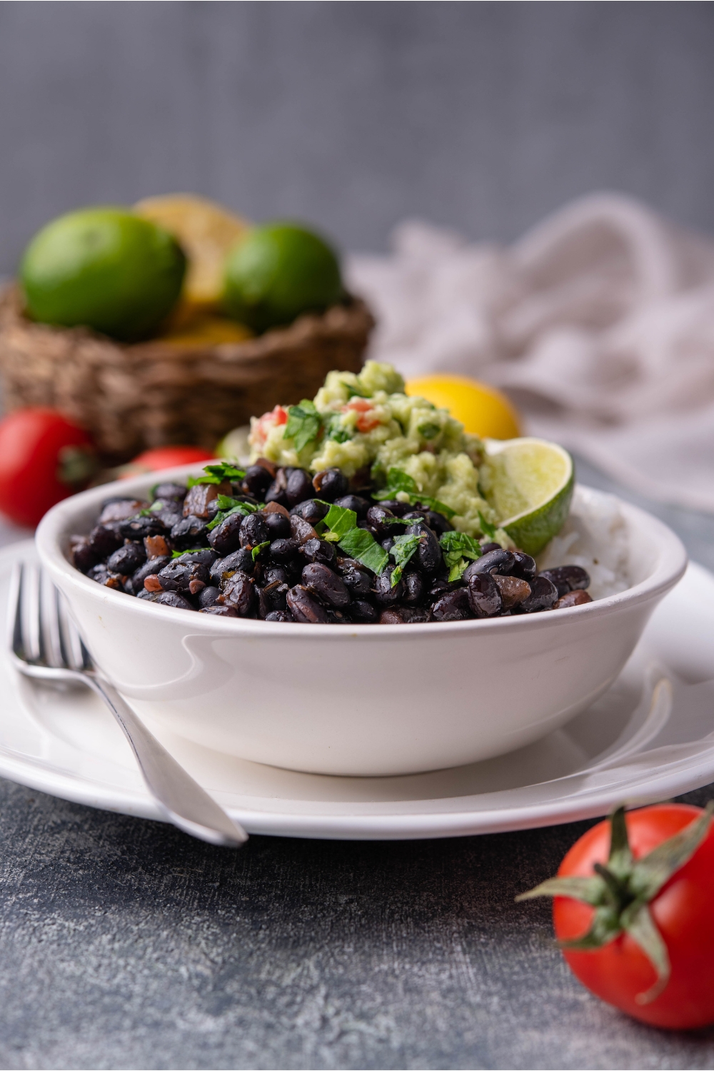 Chipotle black beans in a white bowl layered on a white plate, served with white rice, guacamole, a lime wedge, and fresh herbs. A fork is on the plate and a bowl of limes and lemons is in the background.