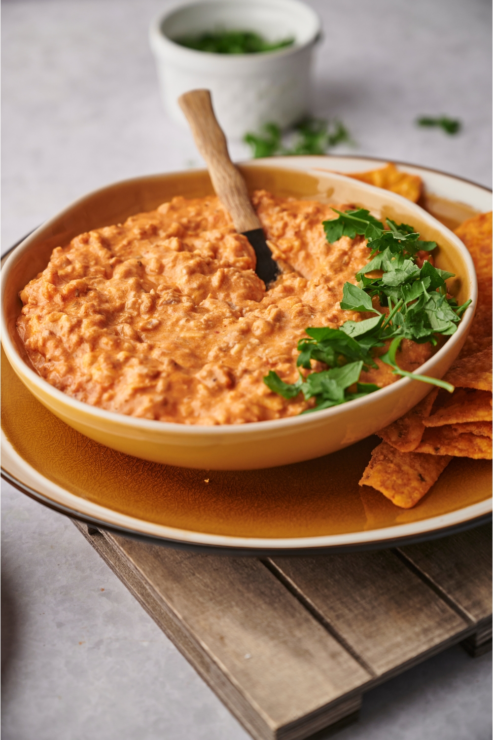 Chili cheese dip in a yellow and white serving bowl with a spoon and fresh herbs in the bowl. The bowl is atop a plate of tortilla chips and a wooden board.